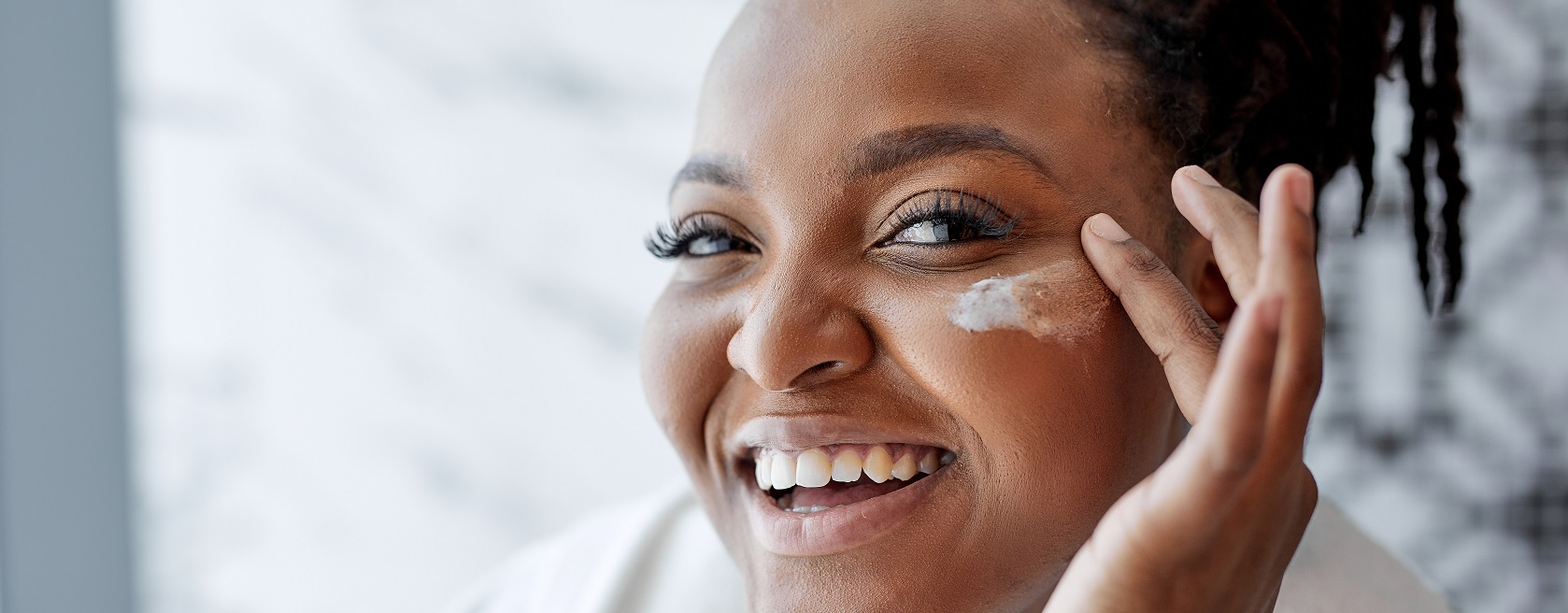 Een vrouw glimlacht terwijl ze crème op haar gezicht aanbrengt.